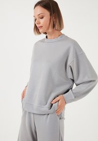 Sudadera gris claro de mangas largas, hecha de una tela suave, con un escote redondeado y bolsillos laterales, de corte holgado y con costuras mínimas.