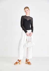 Black long-sleeve top with sheer accents, white high-waisted trousers, brown platform sandals, and a curved white handbag.