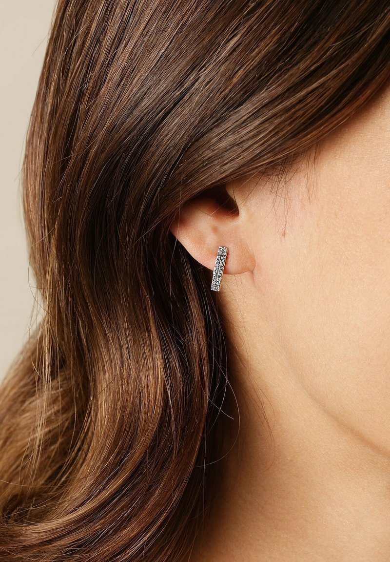 Rechteckige Ohrringe in Silberoptik mit einer Reihe von kleinen klaren Strasssteinen, die vor einem Hintergrund aus glatter Haut und Haaren zur Geltung kommen.