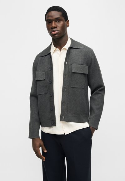 Μαύρος άντρας που φοράει σκούρο γκρι σακάκι με κουμπιά και δύο τσέπες με καπάκια πάνω από λευκό πουκάμισο και μαύρο παντελόνι, σε μονόχρωμο φόντο.