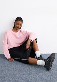 Rosa sweatshirt med rund halsringning och liten logotyp, matchad med en svart knä-lång kjol. Svarta sportskor och vita ankelsockor.