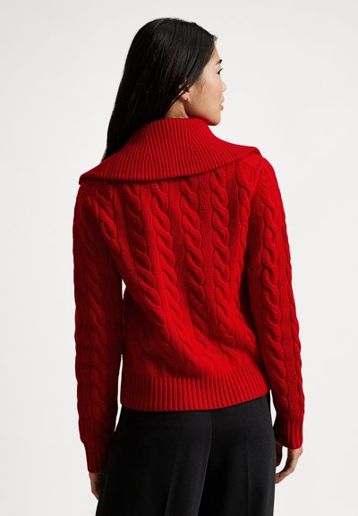 Czerwony sweter z dzianiny z prążkowanym kołnierzem, ozdobiony wzorami warkoczy i dopasowanym dołem. Widać plecy modelki z długimi włosami.