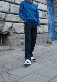 Felpa blu con tasca frontale, pantaloni neri e sneakers bianche con dettagli neri. Sullo sfondo di un muro di pietra e una porta blu.