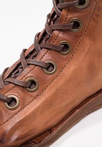 Hnědá kožená kotníková bota s bronzovými kovovými očky a hnědými tkaničkami. Disponuje texturovaným povrchem a mírně zakřiveným podpatkem.