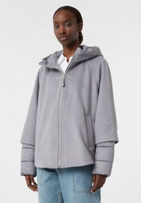 Woman wearing light gray zip-up jacket with puffer sleeves and hood, paired with light blue jeans, against plain white background.