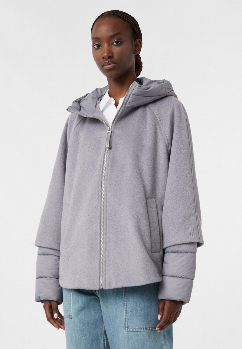 Woman wearing light gray zip-up jacket with puffer sleeves and hood, paired with light blue jeans, against plain white background.