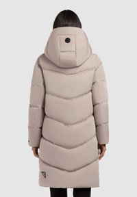 Garšs, gaiši bežs pūkata mētelis ar ševronu kvadrātu rakstu, augstu apkakli un melnu logotipa uz līkuma pie apakšmala. Iespējas ietilpst liela kapuce.