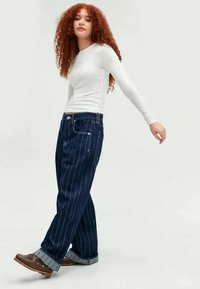 Mulher com cabelo encaracolado vermelho, vestindo uma blusa branca de mangas longas, calças jeans largas azul-escuras com riscas e sapatos castanhos, caminhando para frente.