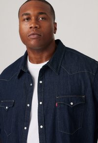Cămașă din denim de culoare albastru închis, cu o textură moale. Prezintă o yoke în stil vestic, închidere cu capse și două buzunare pe piept. Tricou alb vizibil pe dedesubt.