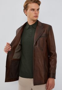 Chaqueta de cuero marrón con solapa muesca, bolsillos frontales y detalle de cremallera. Lleva puesta sobre una camisa polo verde. Textura suave, ajuste entallado.