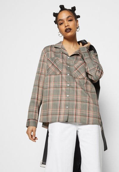 Camisa de flanela xadrez com um padrão verde, vermelho e branco, mangas longas, dois bolsos frontais e um corte reto com a parte de trás ligeiramente mais longa.