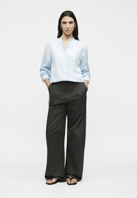 Γυναίκα στέκεται με τα χέρια στις τσέπες, φορά ανοιχτό μπλε πουκάμισο με κουμπιά, φαρδιά μαύρη παντελόνια και μαύρα σανδάλια τύπου φλιπ-φλοπ σε λευκό φόντο.