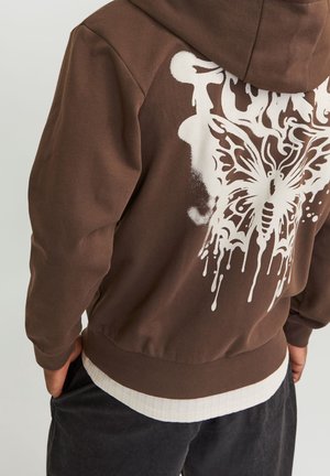 Personne portant un sweat à capuche marron avec un motif de papillon blanc dégoulinant dans le dos, associé à un pantalon noir et une chemise blanche en dessous.