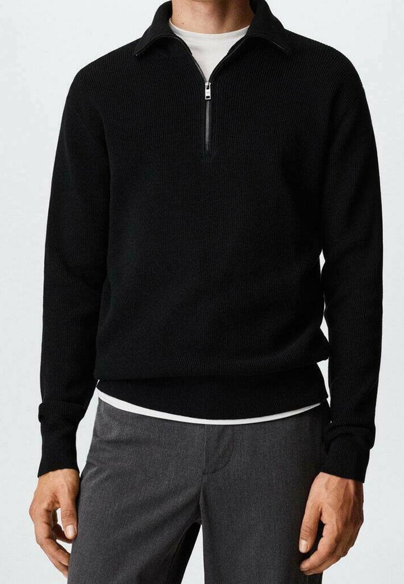 Schwarzer Strickpullover mit einem Viertel-Reißverschluss am Kragen, rippenstruktur und langen Ärmeln. Über einem weißen Rundhalsausschnitt getragen, kombiniert mit grauer Hose.