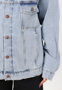 Svjetloplava traper jakna s opuštenim krojem, s smešnim metalnim dugmadi, dva dzepa na grudima i blago pohabanim rubom.