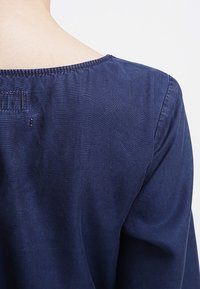 Indumento blu navy con una texture liscia, caratterizzato da un ampio scollo rotondo e maniche lunghe. Panoramica sui dettagli di cucitura sul retro.
