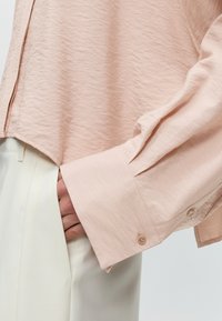 Gros plan d'une personne portant une chemise rose clair à manches longues avec des poignets boutonnés et un pantalon blanc taille haute, main dans la poche.