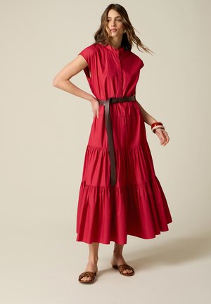 Kvinde iført ærmeløs rød lagdelt maxikjole med brunt bælte, brune armbånd og brune sandaler, stående på beige baggrund.