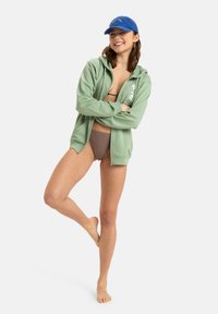 Grön zip-up hoodie med dragsko på huvan, sidofickor och blomstertryckdetaljer, kombinerad med en blå keps och bruna bikiniunderdelar.