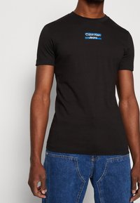 Black cotton t-shirt with a crew neck, short sleeves, and a blue "Calvin Klein Jeans" logo across the chest. Simple design and fit.