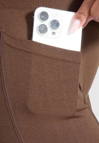 Leggings marroni con una tasca laterale che contiene uno smartphone bianco. Il tessuto è liscio con una leggera lucentezza e presenta dettagli di cucitura visibili.