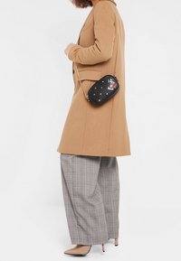 Musta tikattu ristiinkantolaukku kullanvärisillä metalliosilla, jossa on erottuva kuvio ja pitkä olkahihna, käytettynä ruskean takin ja ruudullisten housujen kanssa.