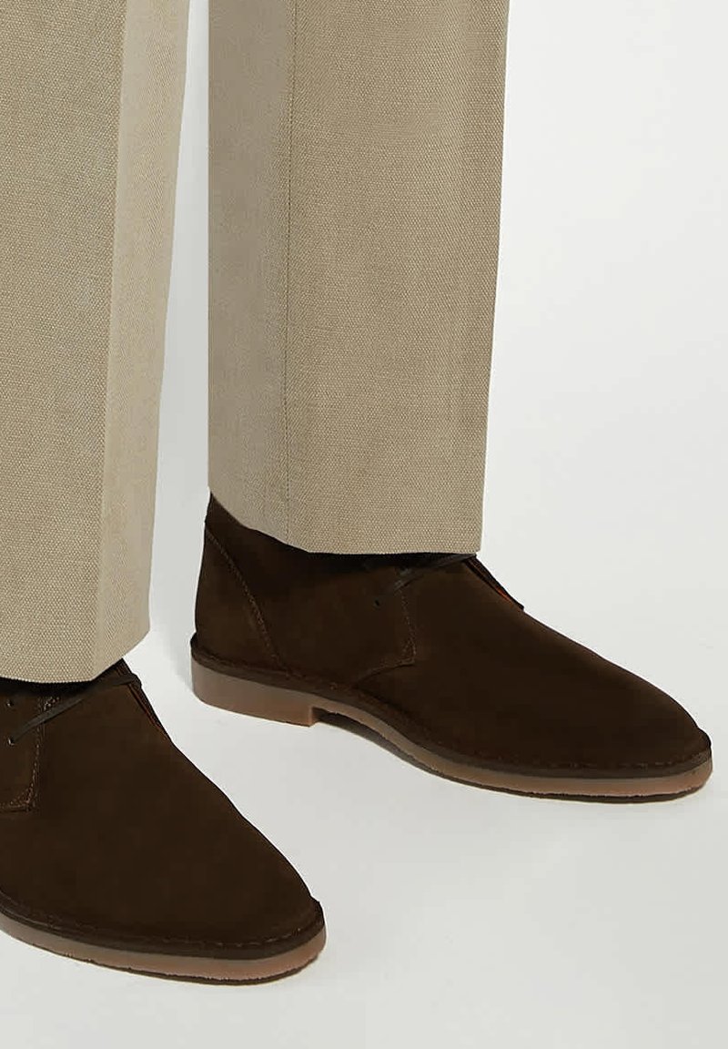Bottes montantes en daim brun foncé à lacets, portées avec un pantalon beige sur fond blanc.