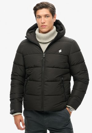 Chaqueta acolchada negra con cierre frontal, capucha y logo blanco. Cuenta con bolsillos laterales y cuello de punto acanalado. Textura suave y diseño estructurado.