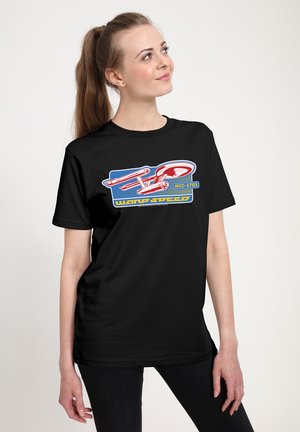 Femme avec une queue de cheval portant un T-shirt noir avec un graphisme de vaisseau spatial rouge et blanc portant l’inscription "NCC-1701" et "Warp Speed."
