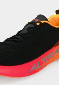 Chaussure de course noire avec une doublure intérieure et une semelle orange néon, logo gris "ALTRA" sur le côté et lacets noirs sur fond blanc.