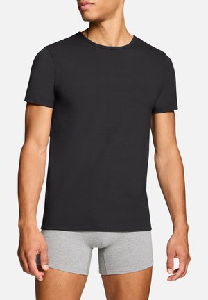 Mann trägt ein eng anliegendes, schlichtes schwarzes T-Shirt mit kurzen Ärmeln und hellgraue Boxer-Shorts, steht vor einem weißen, einfachen Hintergrund.