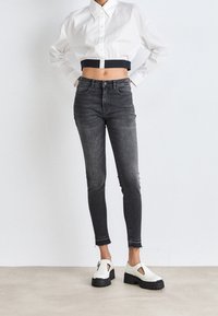 Camicia bianca corta con bottoni e maniche lunghe, abbinata a jeans skinny neri a vita alta e scarpe slip-on bianche con plateau.