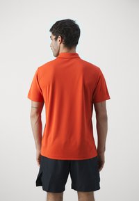 Oransje polo skjorte med krage og korte ermer, laget av et glatt stoff. Bært med svarte shortser, sett fra baksiden.