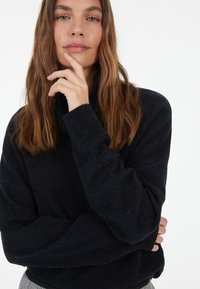 Pullover sweater in dark navy, featuring a high collar and ribbed cuff details, made of soft, textured fabric. Model in a casual pose.