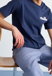 Námořnická modrá bavlněná tričko s kulatým výstřihem, které má bílé logo na levé straně hrudi. Model sedí na dřevěné židli a má na sobě světle šedé kalhoty.