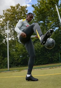 Sportoló illeszkedő szürke hosszú ujjú felsőben és fekete nadrágban, egy szürke focilabdával végrehajt egy ügyességi gyakorlatot, miközben fekete cipőt visel egy zöld mezőn.