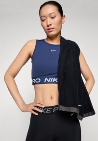 Mørkeblå sports-bh med hvit Nike-logo, svarte leggings med mønstret midjebånd, holder et svart håndkle. Glatt stoff, tettsittende design.