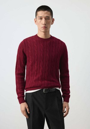 Homme portant un pull en maille torsadée bordeaux et un pantalon noir, debout devant un fond clair uni avec une expression neutre.