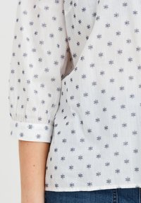 Blusa bianca con un motivo a quadri blu navy. Il tessuto è leggermente trasparente, con maniche arricciate e un orlo arrotondato.