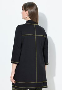 Vrouw met steil bruin haar draagt een zwarte tuniek met lange mouwen, versierd met gele contraststiksel op de rug en manchetten, staand tegen een witte muur.