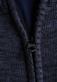 Mörkblå stickad cardigan med ett texturerat mönster och en metalldragkedja med logotyp. Visas med ett lager ovanpå en mörkare blå skjorta.