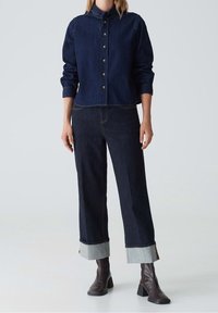 Femme portant une chemise boutonnée en denim noir avec un jean large assorti retroussé aux chevilles et des bottes ankle marron foncé.
