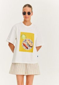 Bílý bavlněný tričko s krátkým rukávem, které na sobě nese žlutý čtvercový grafický motiv talíře s jídlem a modrými vzorovanými okraji.