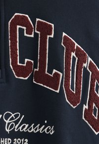 Mörkblå sweatshirt med tjock, vinröd, strukturerad text som står "CLUB" med vit kontur; inkluderar även "Classics" och "Established 2012" i vit brodyr.