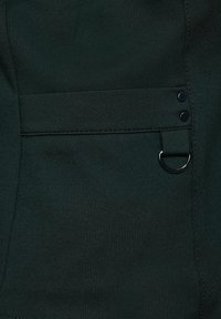 Mörkgrön tyg med en slät textur har en sidoficka, två svarta tryckknappar och en cirkulär metallring som accent.