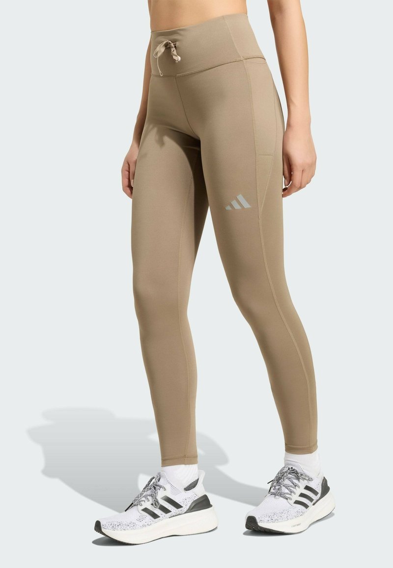 Gros plan sur une personne portant des leggings beige taille haute avec une poche latérale et des chaussures de running claires avec des rayures noires.