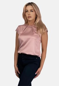 Jasnoróżowa bluzka z krótkim rękawem, okrągłym dekoltem i plisowanym detalem z przodu, zestawiona z ciemnymi spodniami, na białym tle.