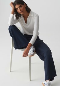 Witte blouse met lange mouwen en een kraag, marineblauwe wijde broek en witte sneakers met blauwe en grijze accenten, zittend op een witte stoel.