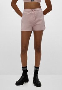 Osoba v pastelově růžových šortkách se stahovací šňůrkou, černých ponožkách s bílým textem a černých robustních botách, stojící na jednoduchém šedém pozadí.