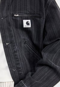 Giacca in denim nero con un motivo a strisce verticali, tasca con zip e un'etichetta bianca con logo. Tessuto testurizzato e design sartoriale.